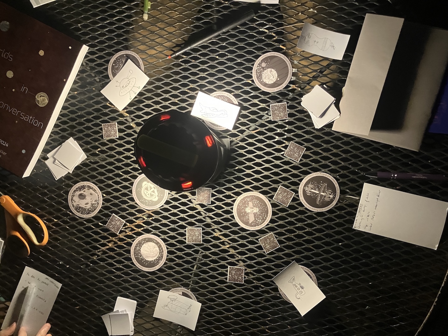 Overhead view of a metal patio table. At its center is a lantern illuminating a surrounding assortment of cardboard tokens representing planets or stations, cut-out squares representing empty space, and drawings of starships on cut-up index cards. There are books, pens, scissors, and other notecards and papers around the edges of the frame.